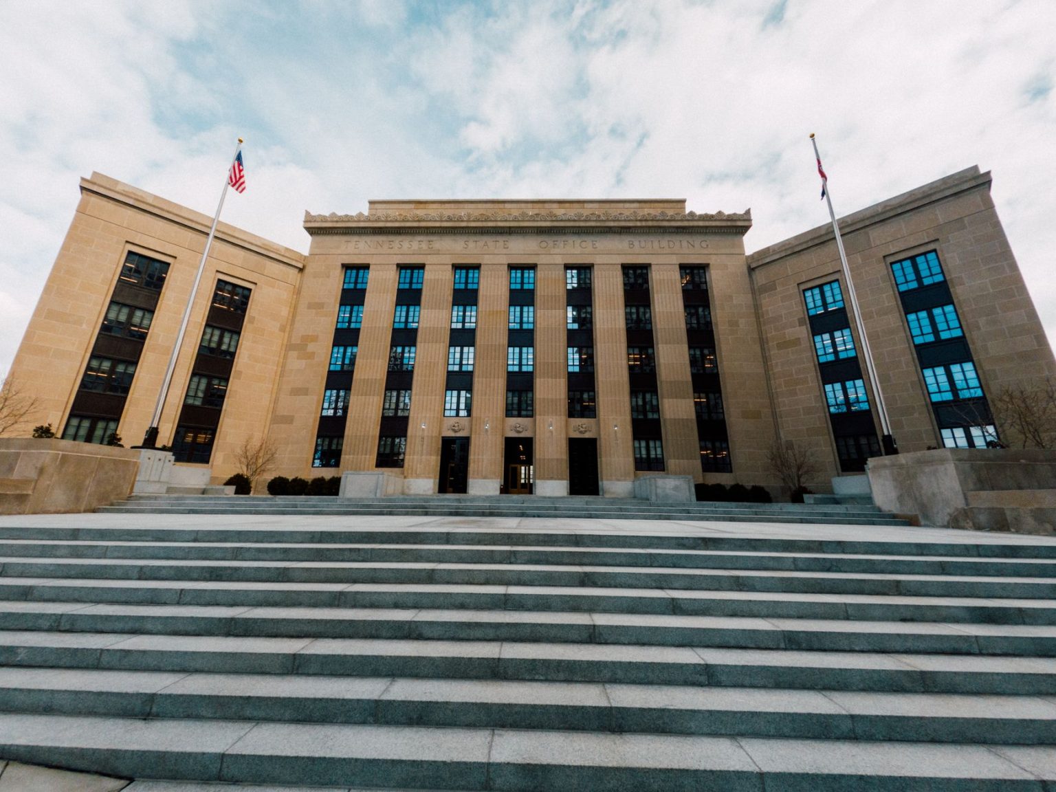 Vista em ângulo baixo do edifício do Federal Reserve, com bandeiras dos Estados Unidos e escadaria sob céu nublado.