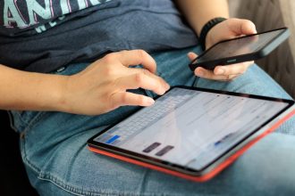 Pessoa usando um tablet e um smartphone em ambiente interno, representando o acesso digital a serviços e informações.