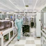 Cientista usando roupa protetora caminha por uma sala limpa de alta tecnologia.