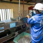 Operário usando macacão azul opera uma mesa vibratória para processamento de minério em uma unidade industrial.
