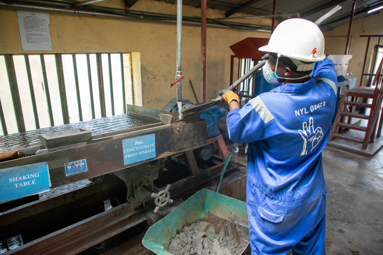 Operário usando macacão azul opera uma mesa vibratória para processamento de minério em uma unidade industrial.