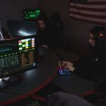 Profissionais analisam sistemas de computador em uma sala pouco iluminada, acompanhando dados relacionados à segurança cibernética.