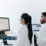 Dois pesquisadores usando jalecos e óculos de proteção analisam dados em um computador dentro de um laboratório.