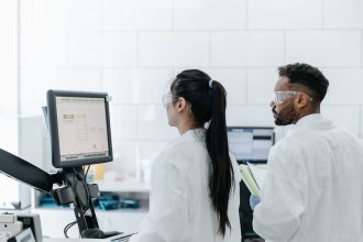 Dois pesquisadores usando jalecos e óculos de proteção analisam dados em um computador dentro de um laboratório.